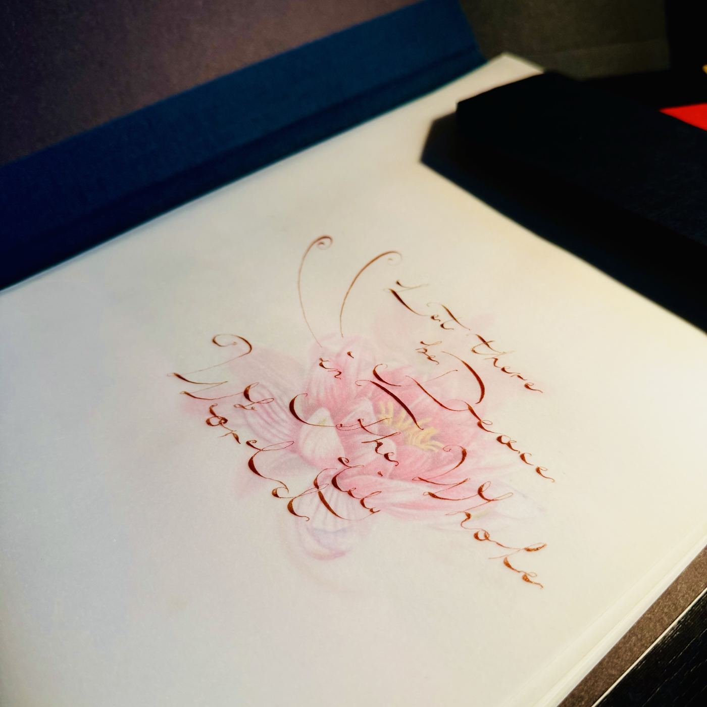 English calligraphy in copper ink reading Let there be peace in the whole world, over a soft pink flower