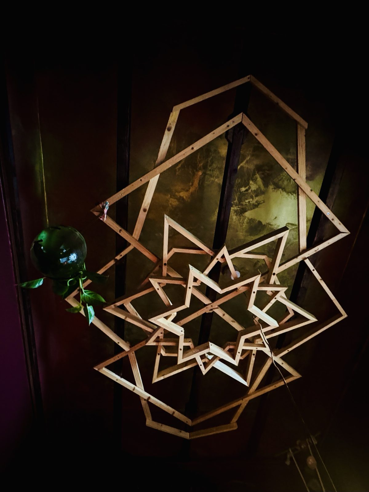 Hand-built structure of light wooden battens fixed to a dark ceiling: rotated squares, larger polygons and star shapes turning within one another, with a trailing plant in a glass globe beside it